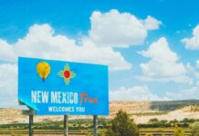 Blue “Welcome to New Mexico” highway sign under bright sky and clouds.