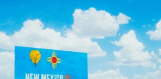 Blue “Welcome to New Mexico” highway sign under bright sky and clouds.