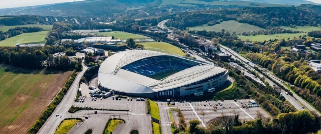 Aerial view of a large stadium with a blue interior, surrounded by parking lots and lush greenery