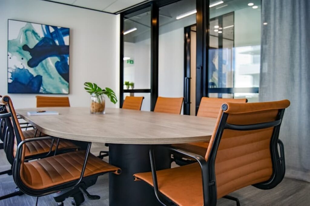 Modern conference room with a wooden table, brown leather chairs, glass walls, and a large abstract painting on the wall.