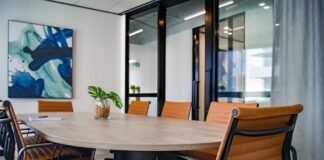 Modern conference room with a wooden table, brown leather chairs, glass walls, and a large abstract painting on the wall.