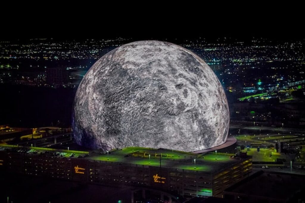 A high-resolution aerial photo of the Las Vegas Sphere lit up as a massive moon, set against the city’s night skyline.