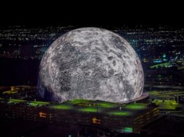 A high-resolution aerial photo of the Las Vegas Sphere lit up as a massive moon, set against the city’s night skyline.