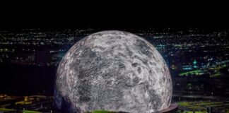 A high-resolution aerial photo of the Las Vegas Sphere lit up as a massive moon, set against the city’s night skyline.