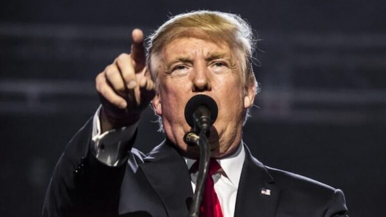 Donald Trump speaking at a rally, pointing forward while addressing the audience from behind a microphone.