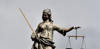 A stone statue of Lady Justice stands against a cloudy sky, holding a sword in one hand and a set of scales in the other.