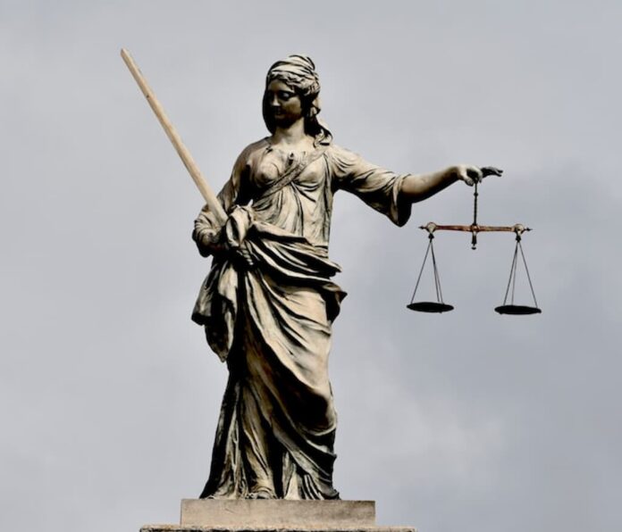 A stone statue of Lady Justice stands against a cloudy sky, holding a sword in one hand and a set of scales in the other.