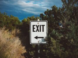 Crypto.com Stops Offering Sports Event Contracts in Arizona, Eight Other States A roadside exit sign with an arrow pointing left, surrounded by trees and brush under a cloudy sky