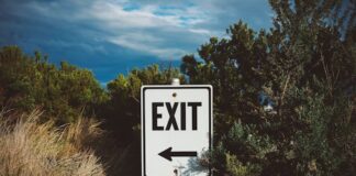 Crypto.com Stops Offering Sports Event Contracts in Arizona, Eight Other States A roadside exit sign with an arrow pointing left, surrounded by trees and brush under a cloudy sky