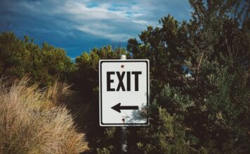A roadside exit sign with an arrow pointing left, surrounded by trees and brush under a cloudy sky