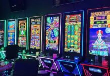 A row of illuminated slot-style gaming machines with colorful screens and chairs lined up in a dark room.