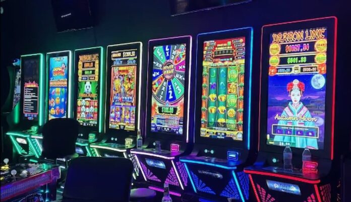 A row of illuminated slot-style gaming machines with colorful screens and chairs lined up in a dark room.