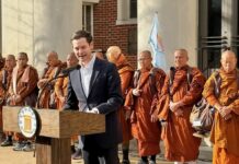 Alabama state Rep. Phillip Ensler speaks at a campaign event outside a government building, standing at a podium with a group of Buddhist monks behind him.