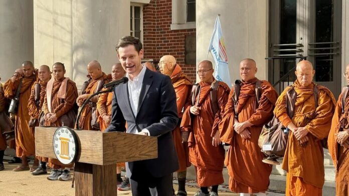 Alabama state Rep. Phillip Ensler speaks at a campaign event outside a government building, standing at a podium with a group of Buddhist monks behind him.