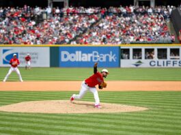 A Cleveland Guardians pitcher delivers a pitch from the mound during a daytime MLB game with a full crowd in the stands.