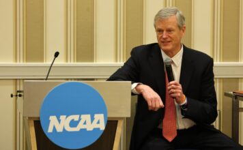NCAA President Charlie Baker speaks into a microphone while seated beside an NCAA-branded podium at a conference event.