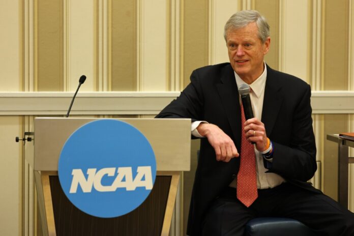 NCAA President Charlie Baker speaks into a microphone while seated beside an NCAA-branded podium at a conference event.