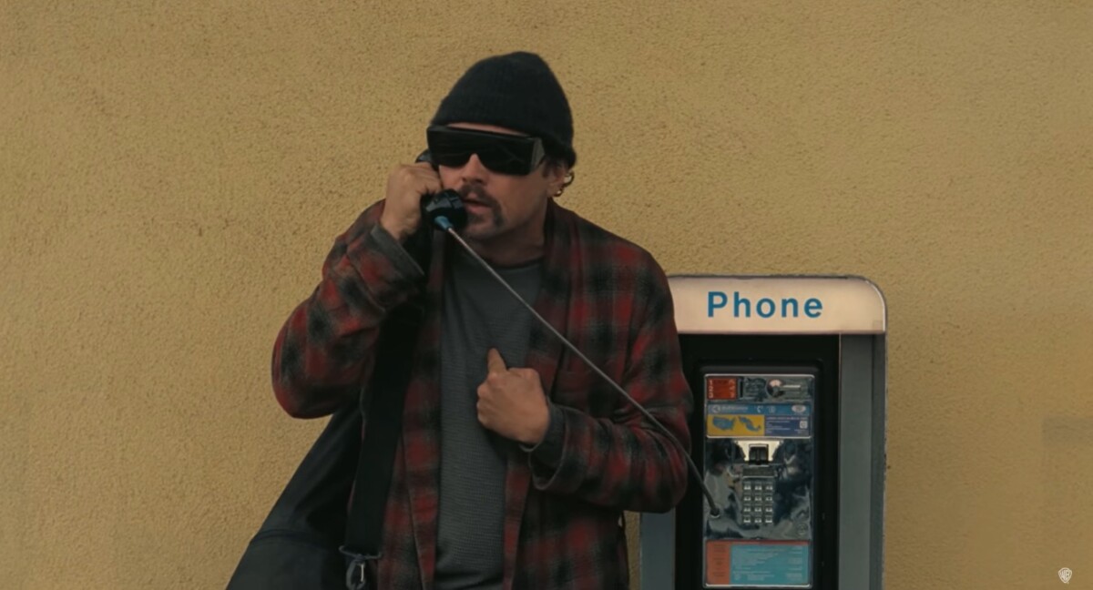 Leonardo DiCaprio wearing big black sunglasses and a cap, standing next to the payphone and speaking on the phone