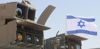 Israel Indicts Two Over Use of Military Secrets for Polymarket Bets Israeli military equipment displayed beneath an Israeli flag during a defense exercise.