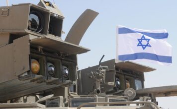 Israel Indicts Two Over Use of Military Secrets for Polymarket Bets Israeli military equipment displayed beneath an Israeli flag during a defense exercise.