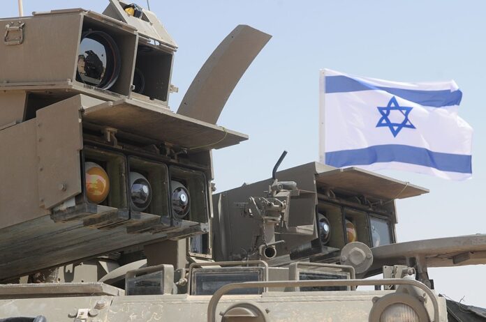 Israel Indicts Two Over Use of Military Secrets for Polymarket Bets Israeli military equipment displayed beneath an Israeli flag during a defense exercise.