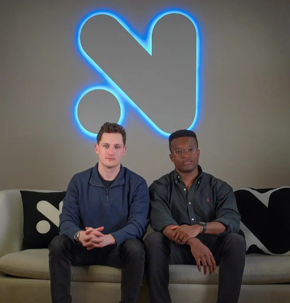 Novig founders seated on a couch beneath the company’s illuminated logo at the firm’s headquarters.