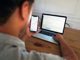 Person holding a smartphone with a trading chart displayed while working on a laptop showing stock market data.