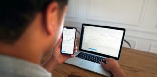 New Poll Finds Americans More Likely to Call Prediction Markets Betting Than Investing Person holding a smartphone with a trading chart displayed while working on a laptop showing stock market data.
