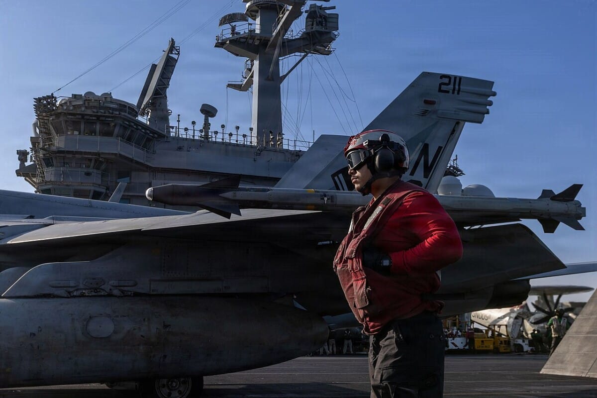 U.S. Navy crew member stands beside a fighter jet on an aircraft carrier flight deck during operations