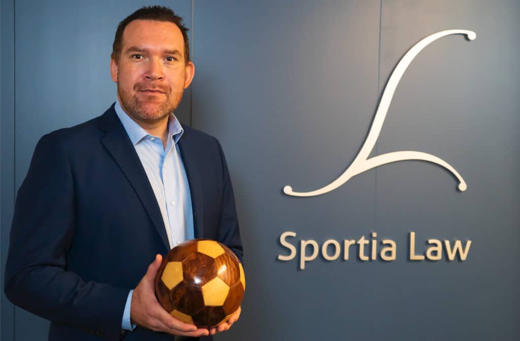 Sports integrity expert Rodrigo Arias Grillo in a navy suit holding a soccer ball in front of the Sportia Law sign and logo.